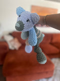 Hand holding a crochet patchwork bear in shades of blue, green, and tan in front of blurred background