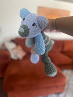 Hand holding a crochet patchwork bear in shades of blue, green, and tan in front of blurred background