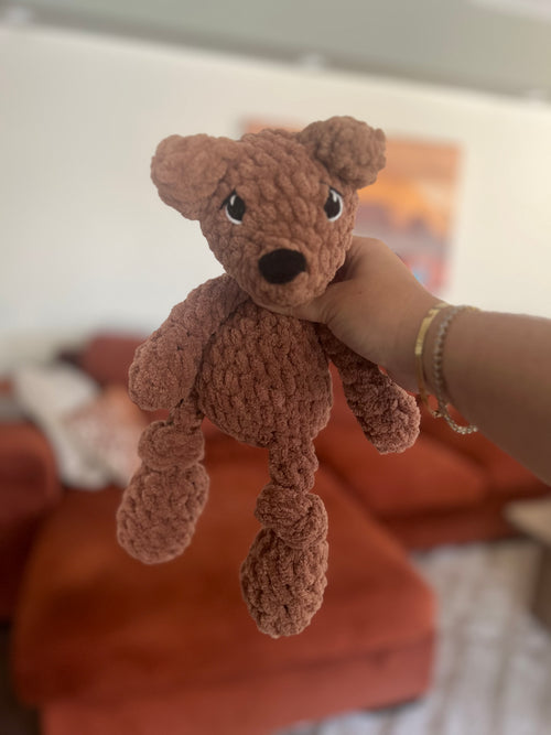 Hand holding a small crochet brown teddy bear against a blurred indoor background