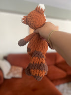Hand holding a small orange, brown, and white crochet red panda toy against a blurred background