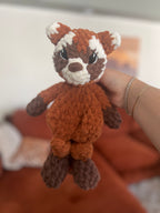 Hand holding a small orange, brown, and white crochet red panda toy against a blurred background