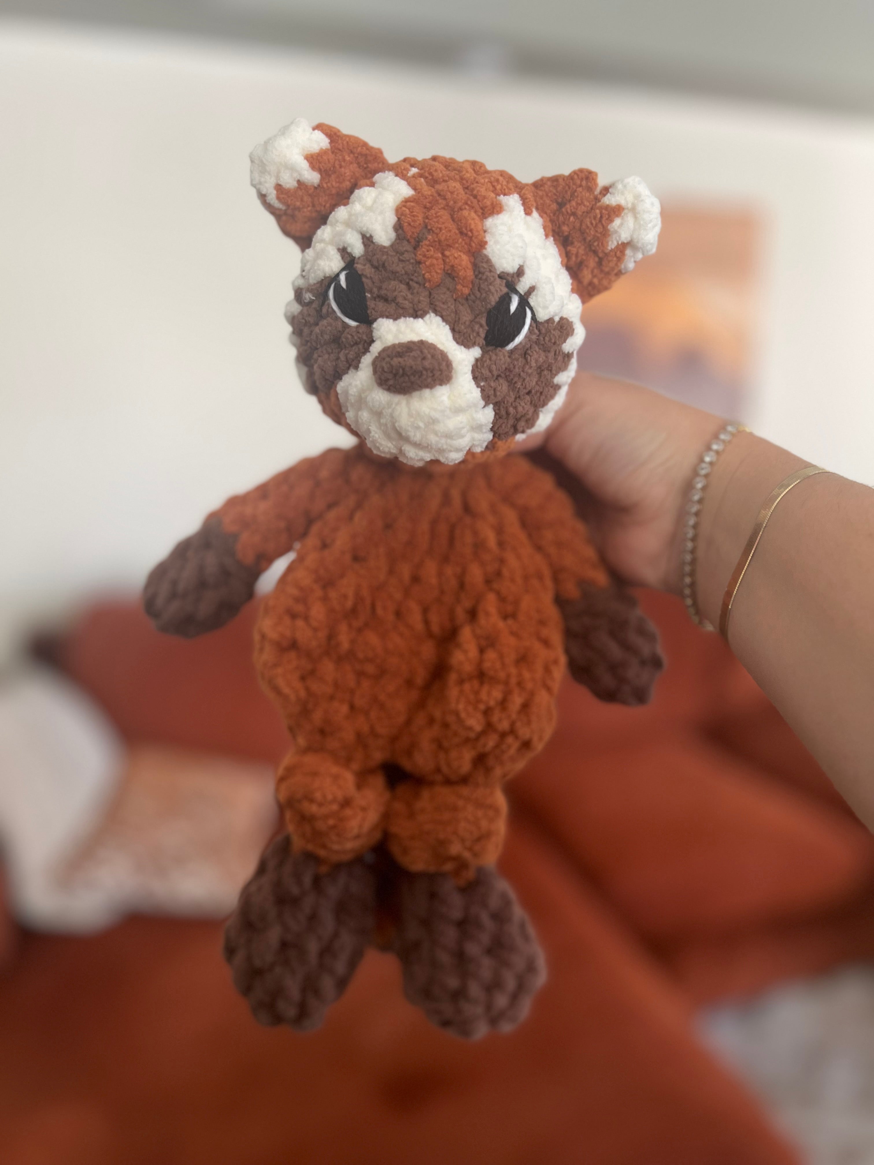 Hand holding a small orange, brown, and white crochet red panda toy against a blurred background