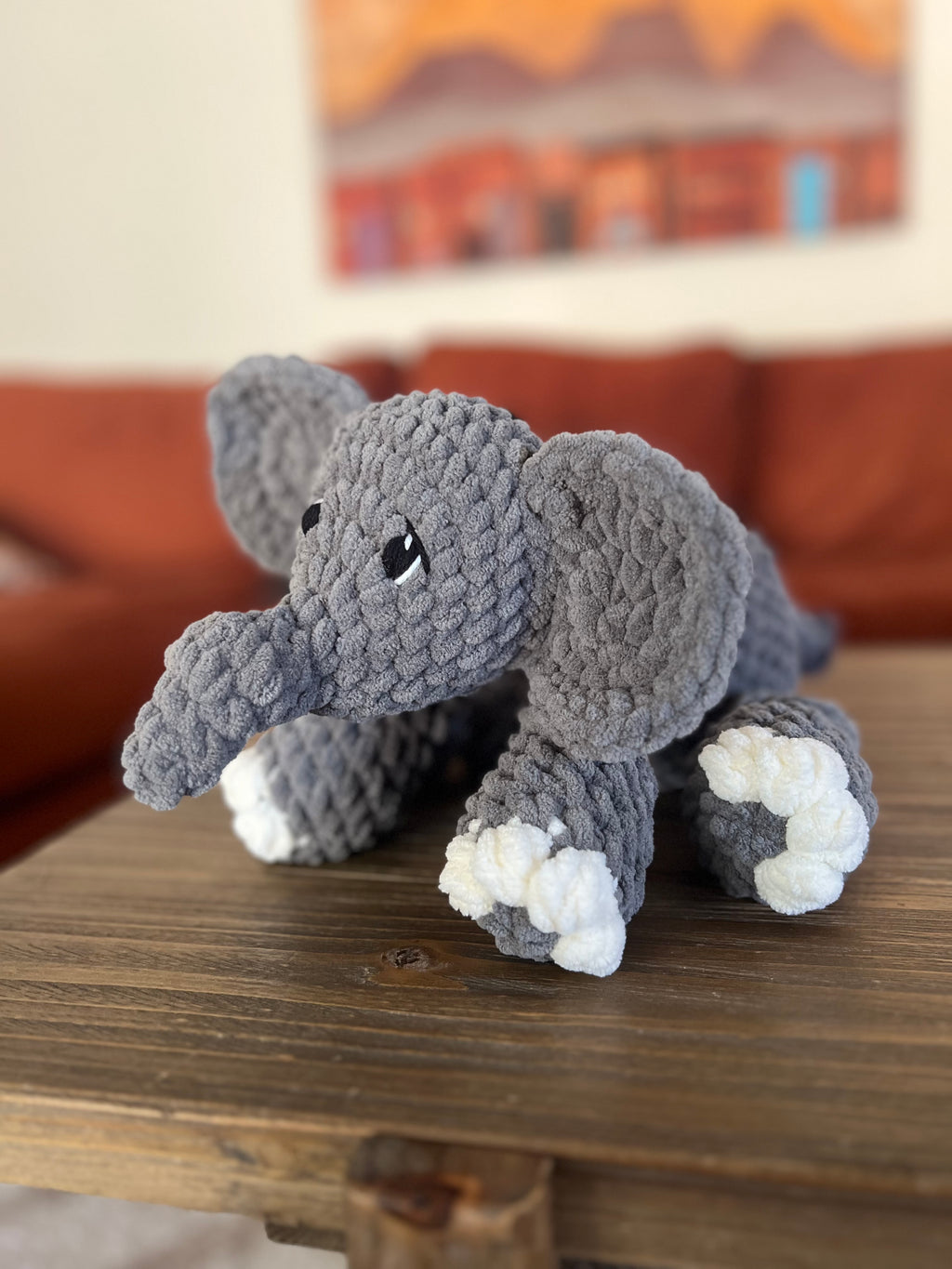 Crochet elephant on wooden table with blurred orange couch and painting in the background