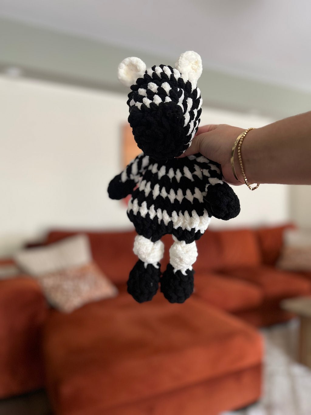 Hand holding a small black and white striped crochet toy zebra in a living room setting.