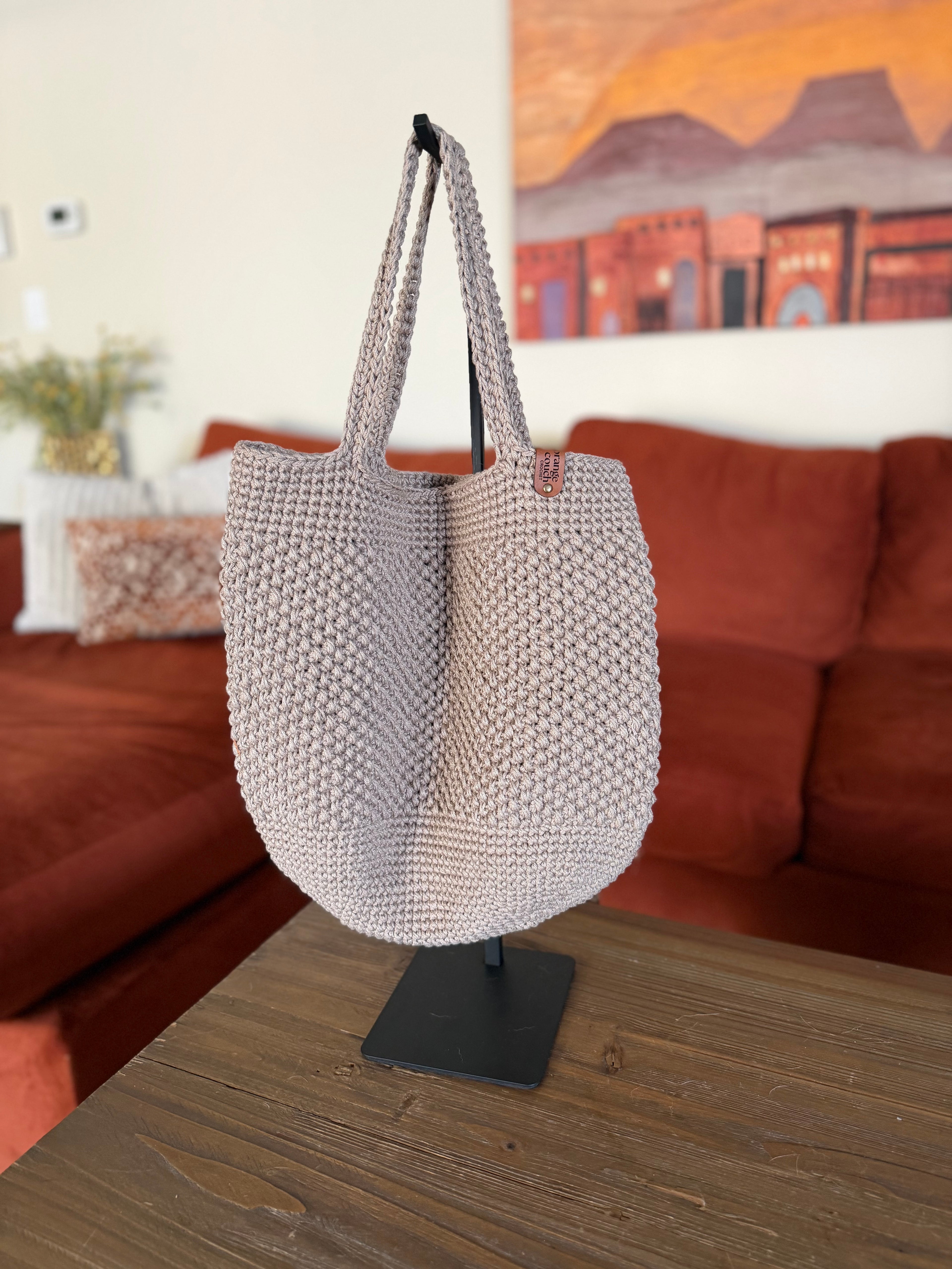 Beige crochet tote bag on a stand in front of a orange couch with a painting in the background