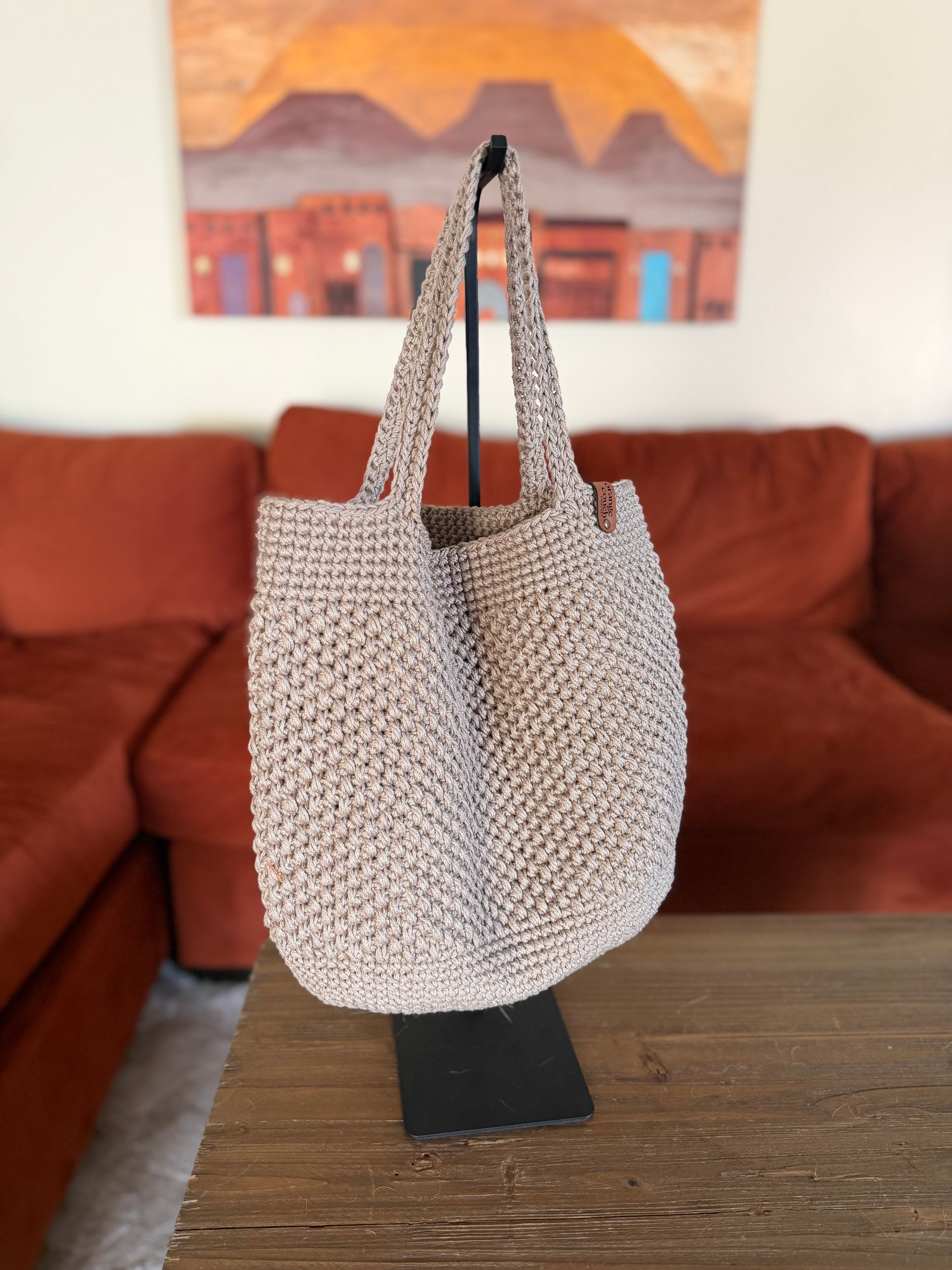 Beige crochet tote bag on a stand in front of a orange couch with a painting in the background