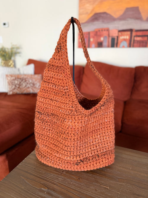 Orange asymmetrical crochet bag on a wooden table with a couch and painting in the background