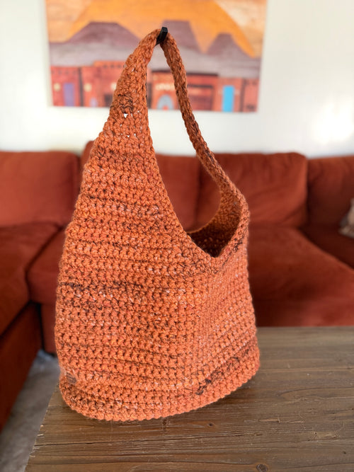 Orange asymmetrical crochet bag on a wooden table with a couch and painting in the background