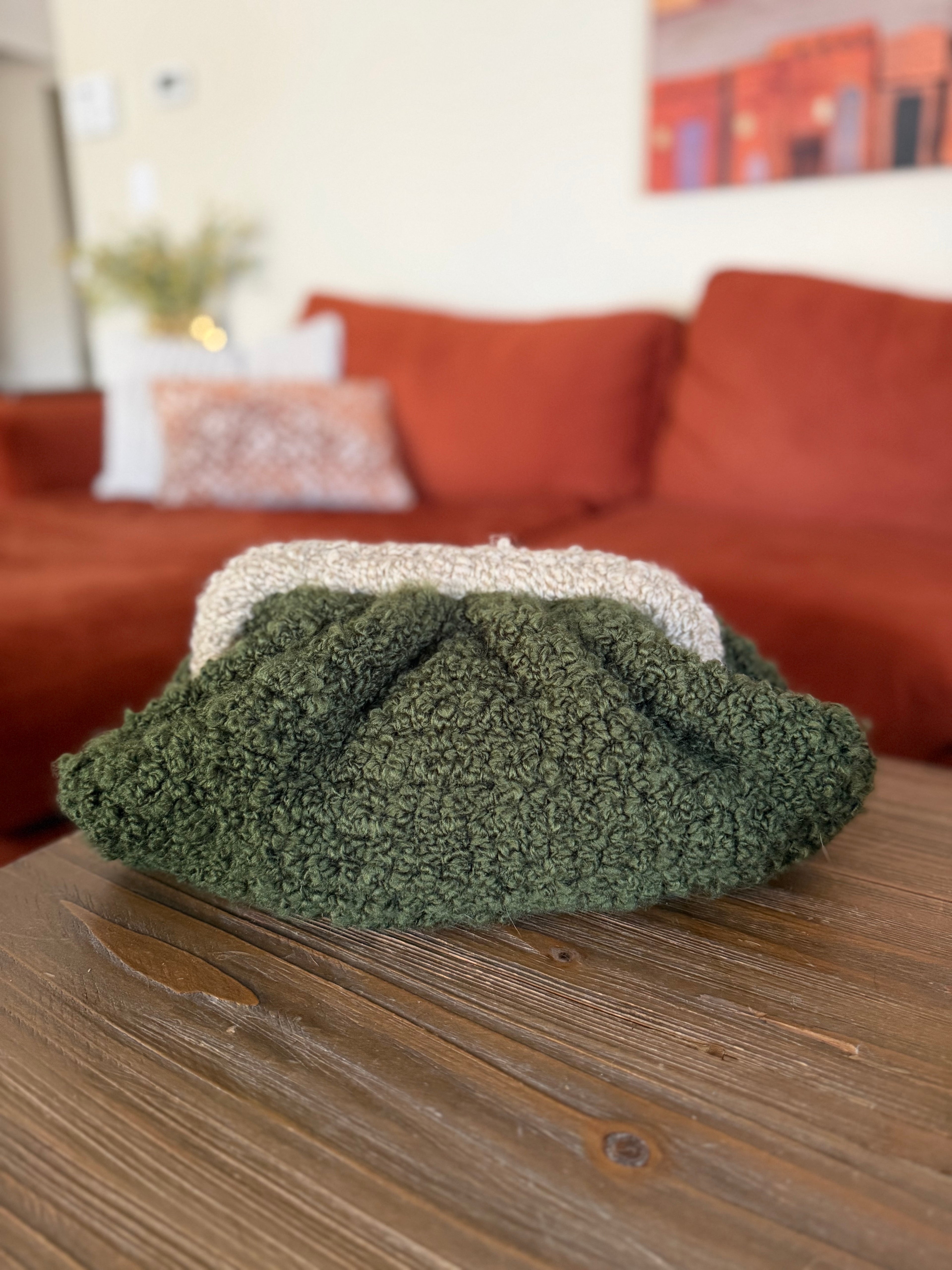 Green crochet clutch on a wooden table with a brown sofa and colorful painting in the background