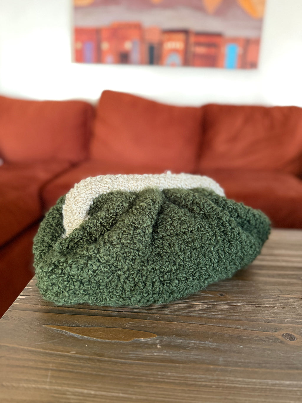 Green crochet clutch on a wooden table with a brown sofa and colorful painting in the background
