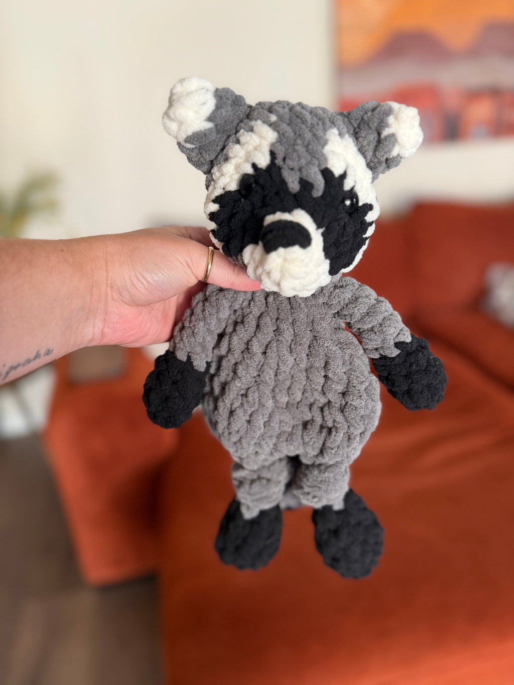 Grey, black, and white crochet raccoon lovey in front of blurred orange couch