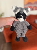 Grey, black, and white crochet raccoon lovey in front of blurred orange couch