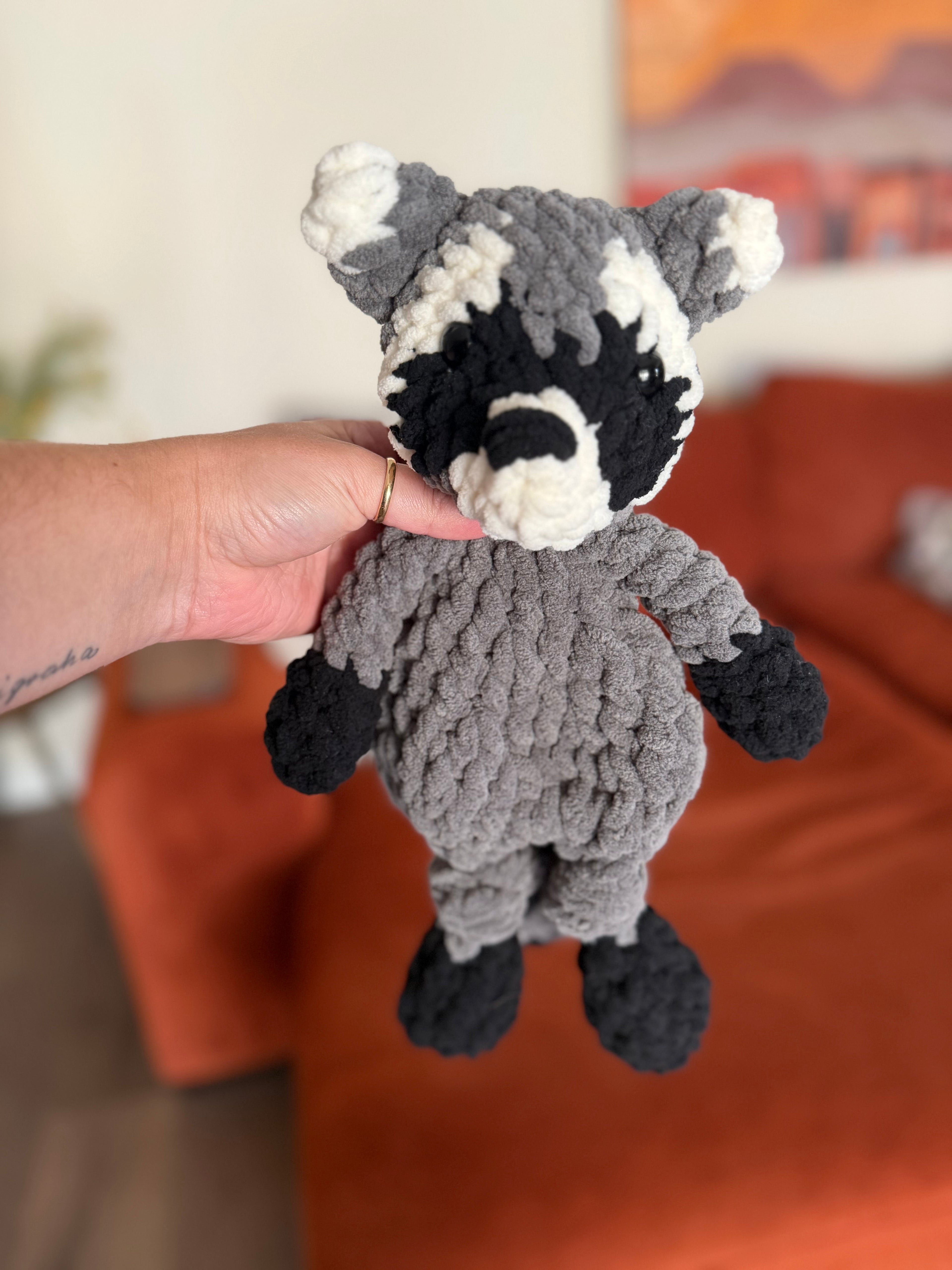 Grey, black, and white crochet raccoon lovey in front of blurred orange couch
