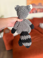 Grey, black, and white crochet raccoon lovey in front of blurred orange couch