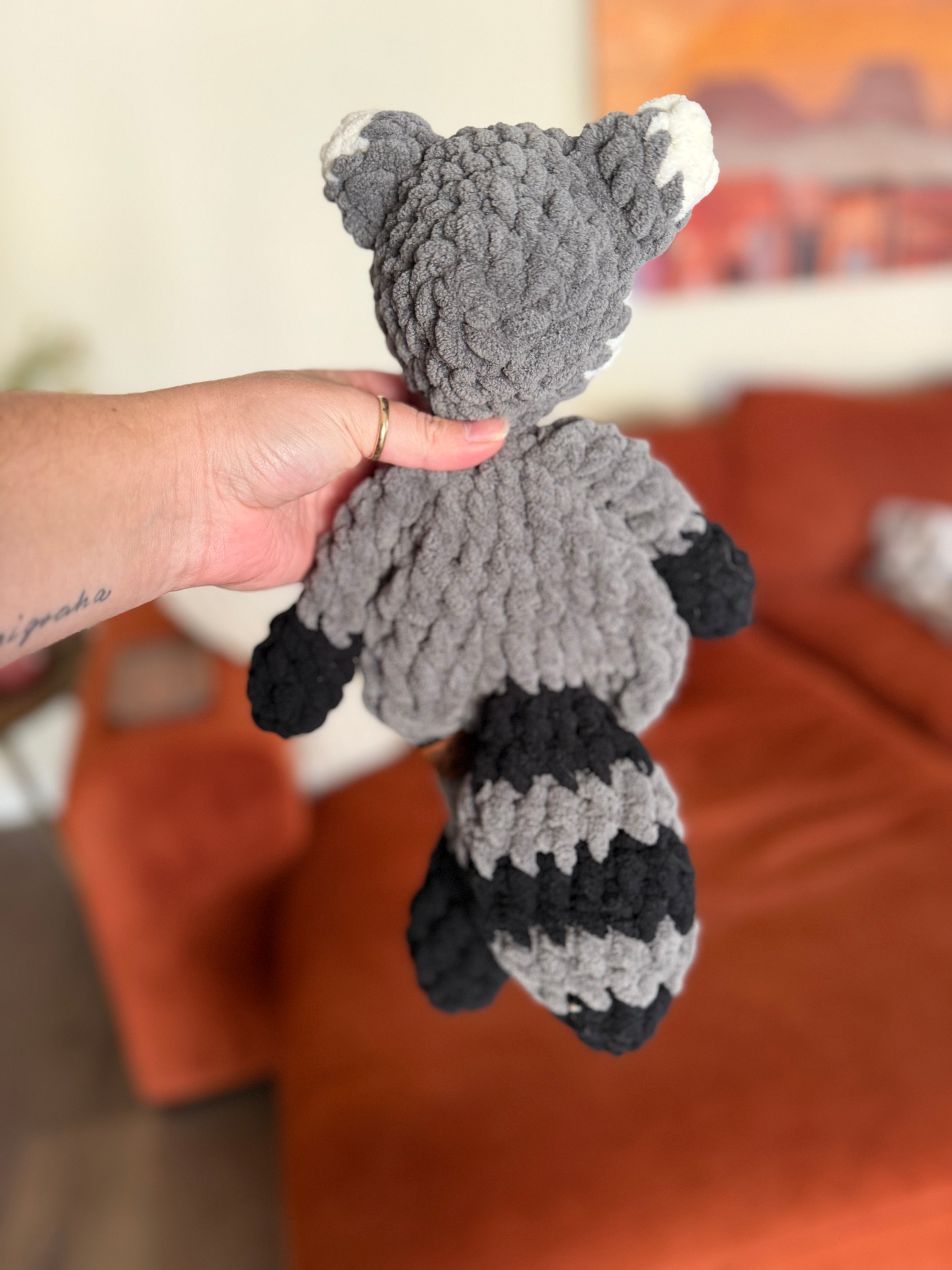 Grey, black, and white crochet raccoon lovey in front of blurred orange couch