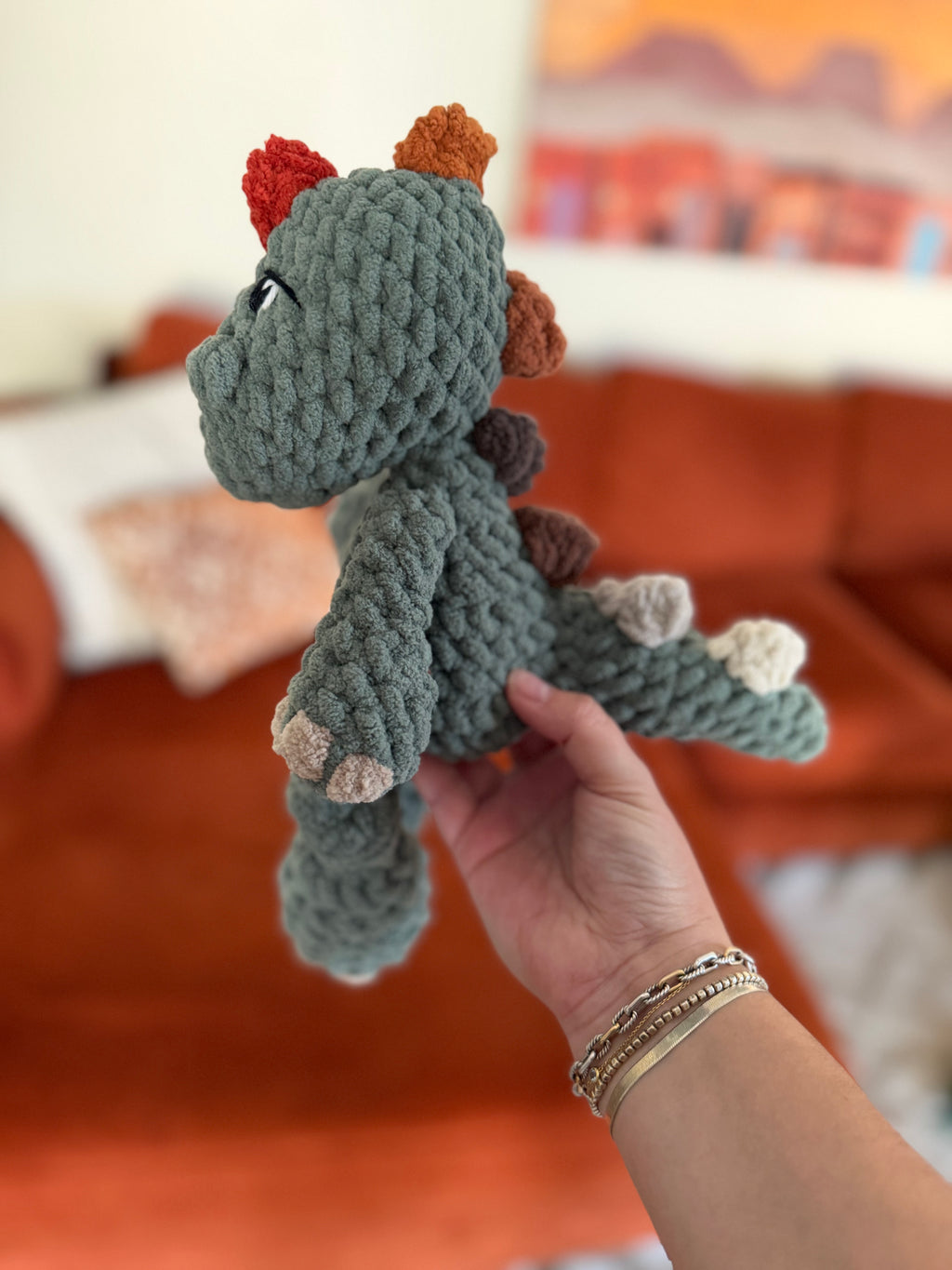 Green crochet dinosaur plushie with ombre spikes beginning in red and ending in cream. Crochet item in front of blurred orange couch.