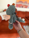 Green crochet dinosaur plushie with ombre spikes beginning in red and ending in cream. Crochet item in front of blurred orange couch.