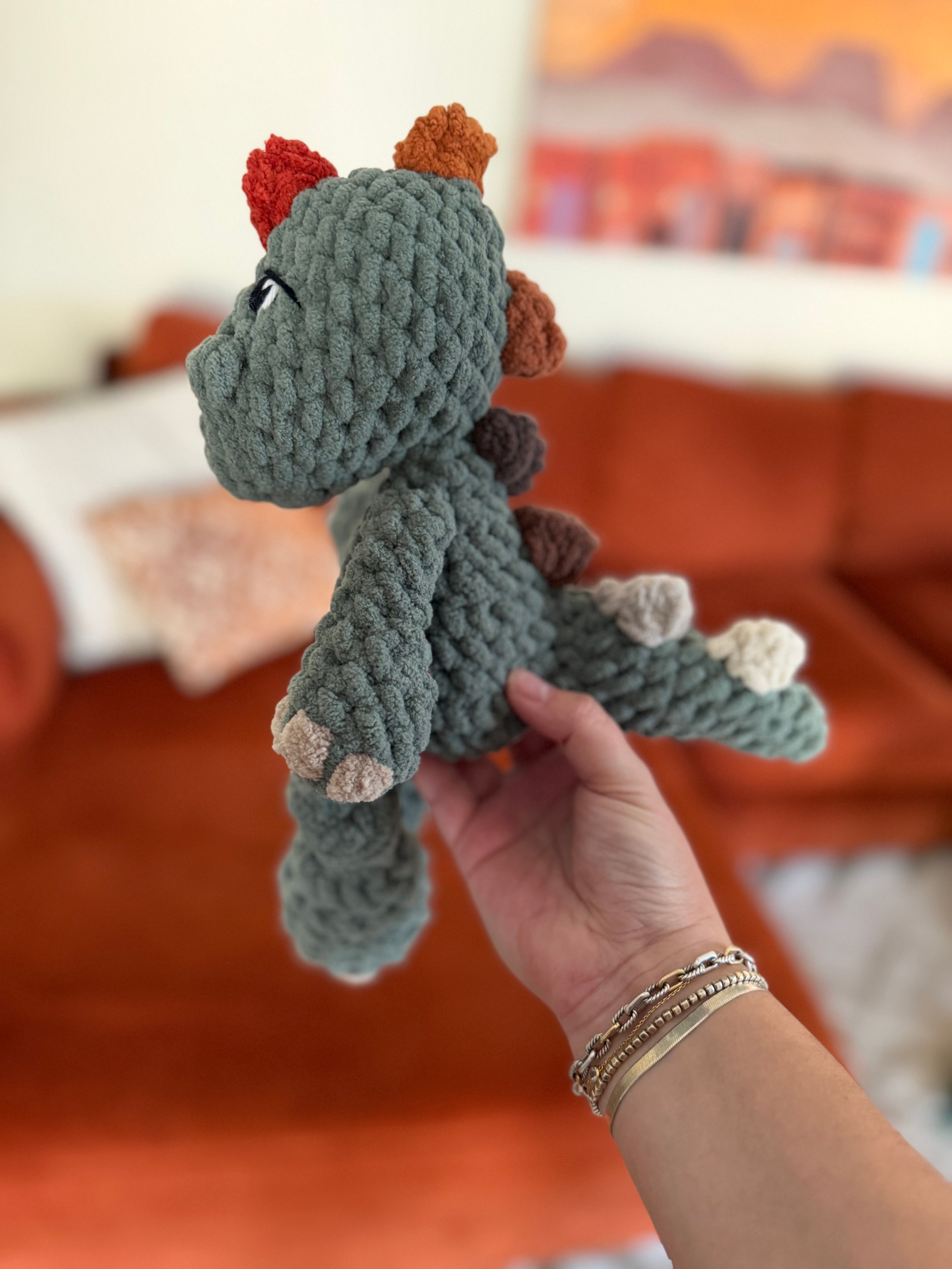 Green crochet dinosaur plushie with ombre spikes beginning in red and ending in cream. Crochet item in front of blurred orange couch.
