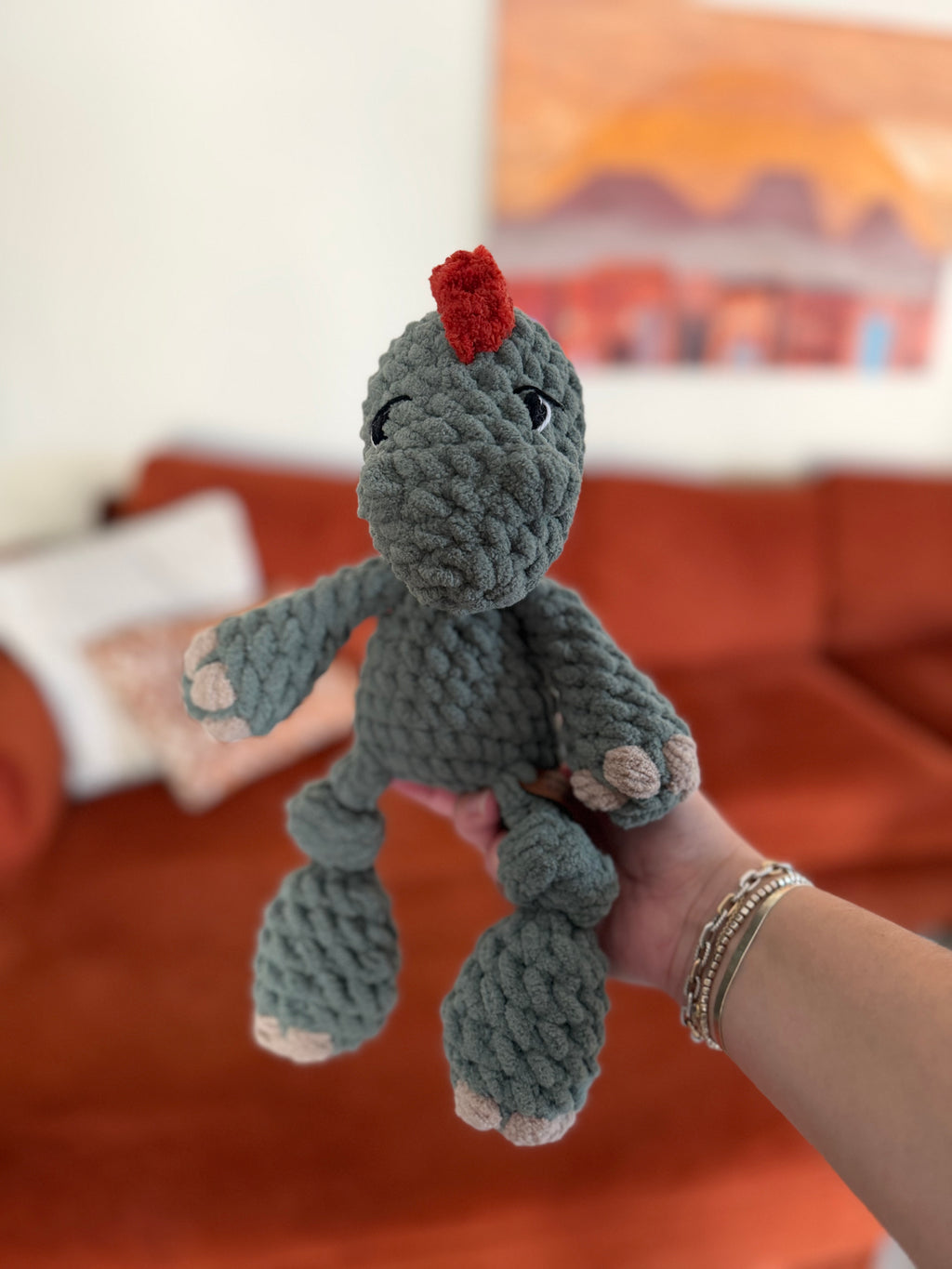 Green crochet dinosaur plushie. Crochet item in front of blurred orange couch.