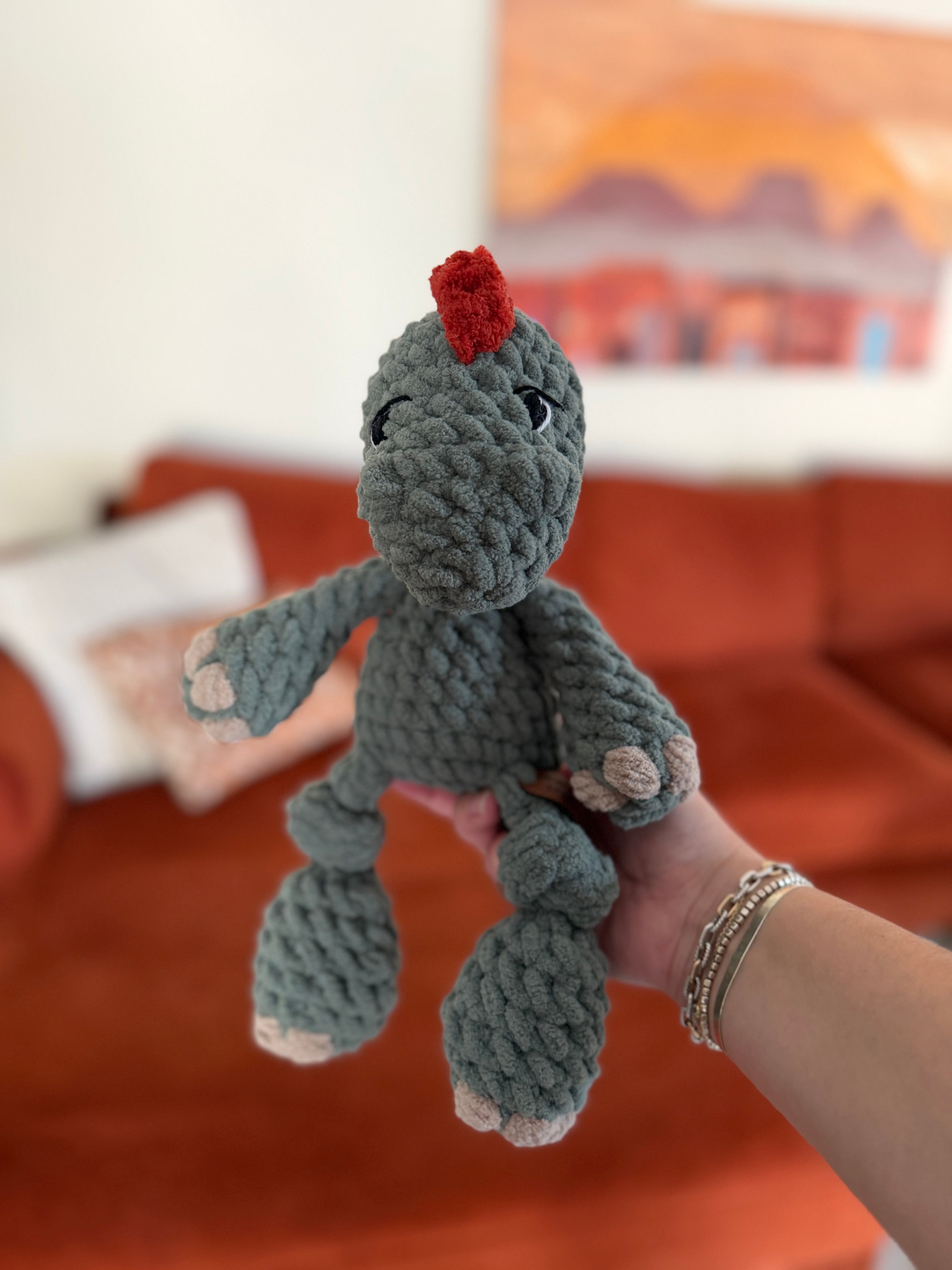 Green crochet dinosaur plushie. Crochet item in front of blurred orange couch.