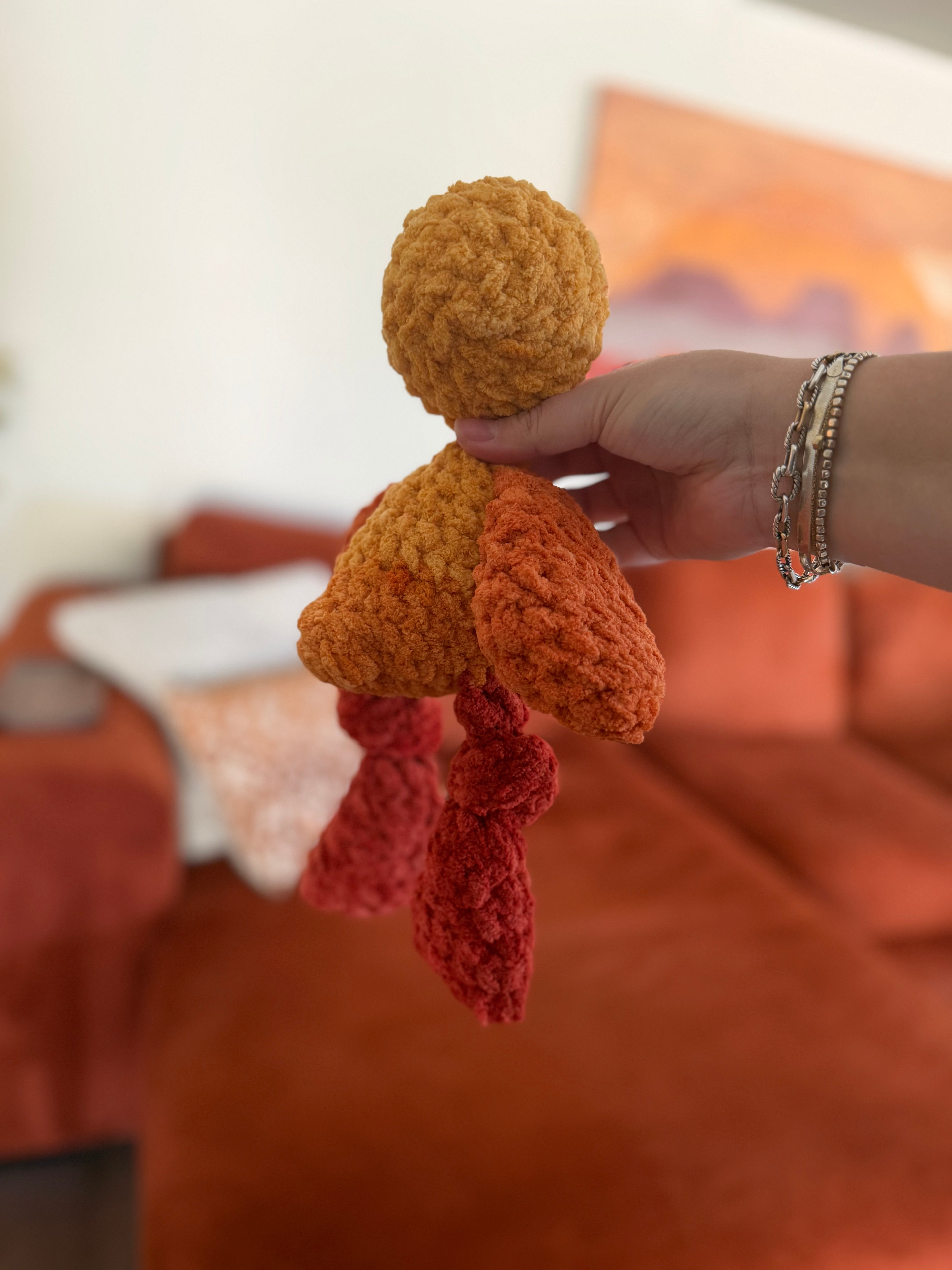 Hand holding a orange, yellow, and red multi-colored crochet chicken plushie against an orange couch