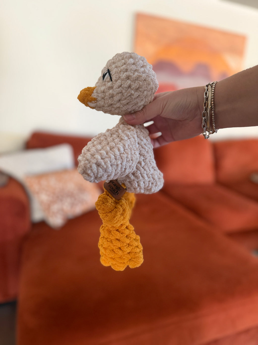 Hand holding a small beige and yellow crochet chicken plushie against a blurred orange couch