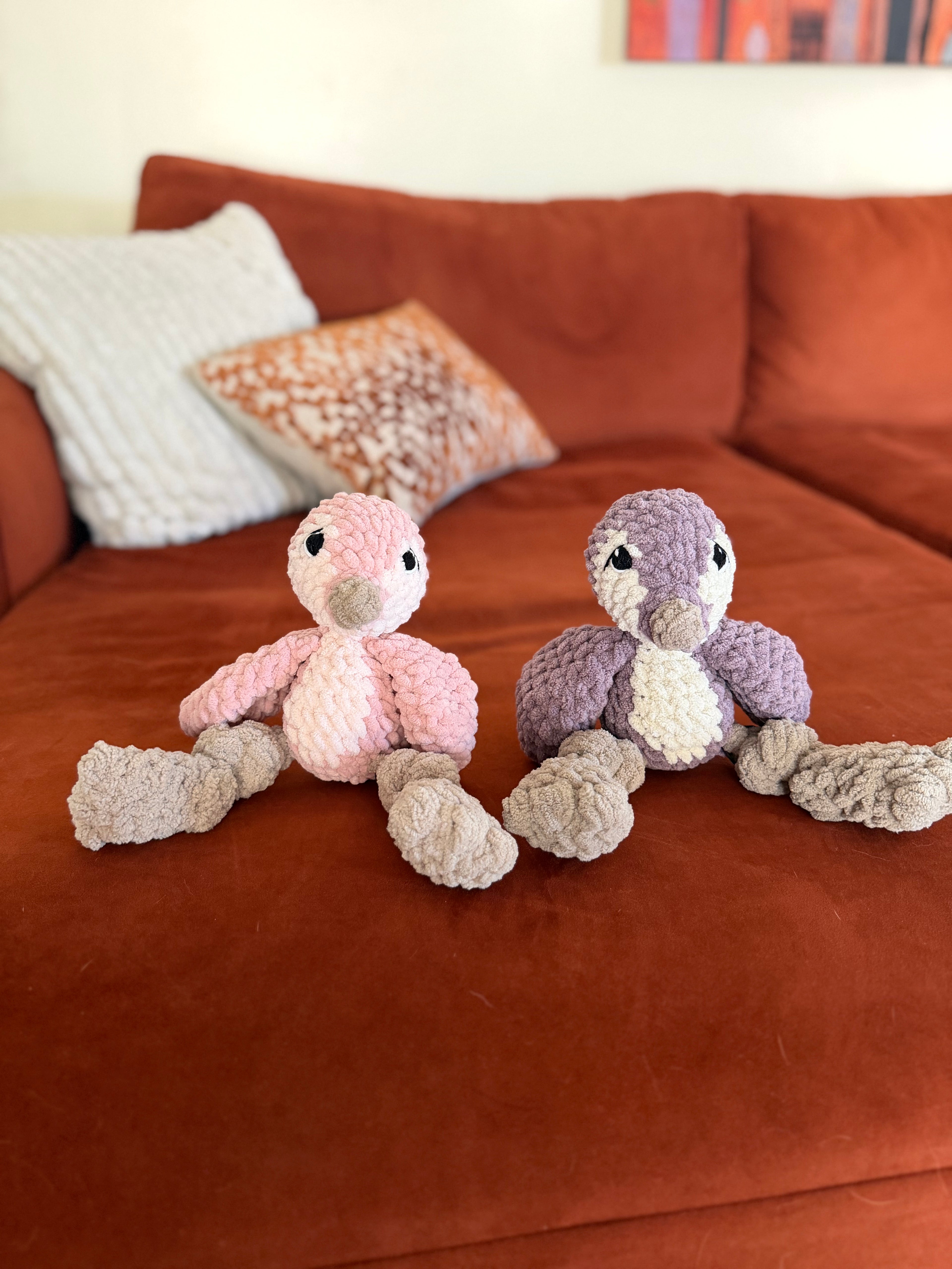 Two crochet penguins on an orange couch. The penguin on the left is pink and white. The penguin on the right is purple and white.