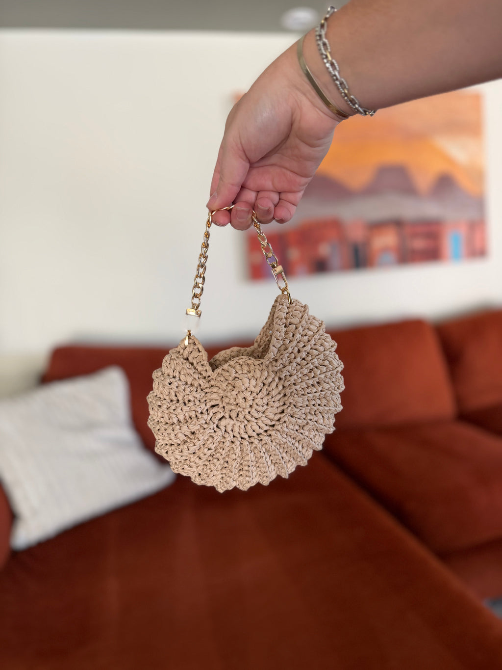 A hand holding a mini shell crochet bag with a gold chain in front of a blurred orange couch