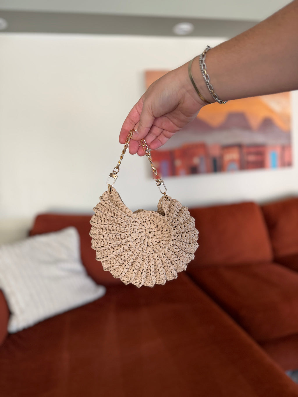 A hand holding a mini shell crochet bag with a gold chain in front of a blurred orange couch