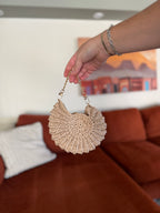 A hand holding a mini shell crochet bag with a gold chain in front of a blurred orange couch