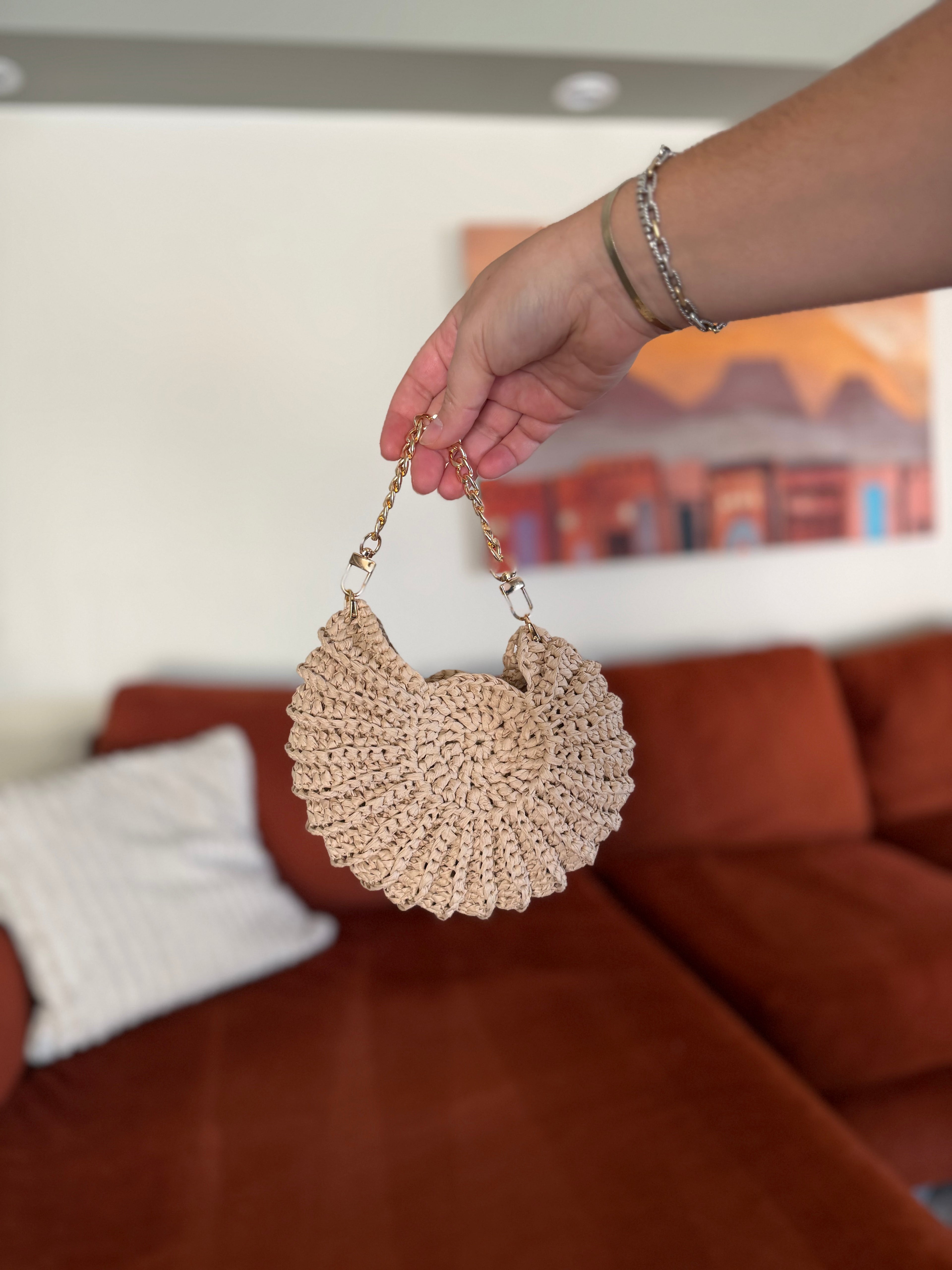 A hand holding a mini shell crochet bag with a gold chain in front of a blurred orange couch