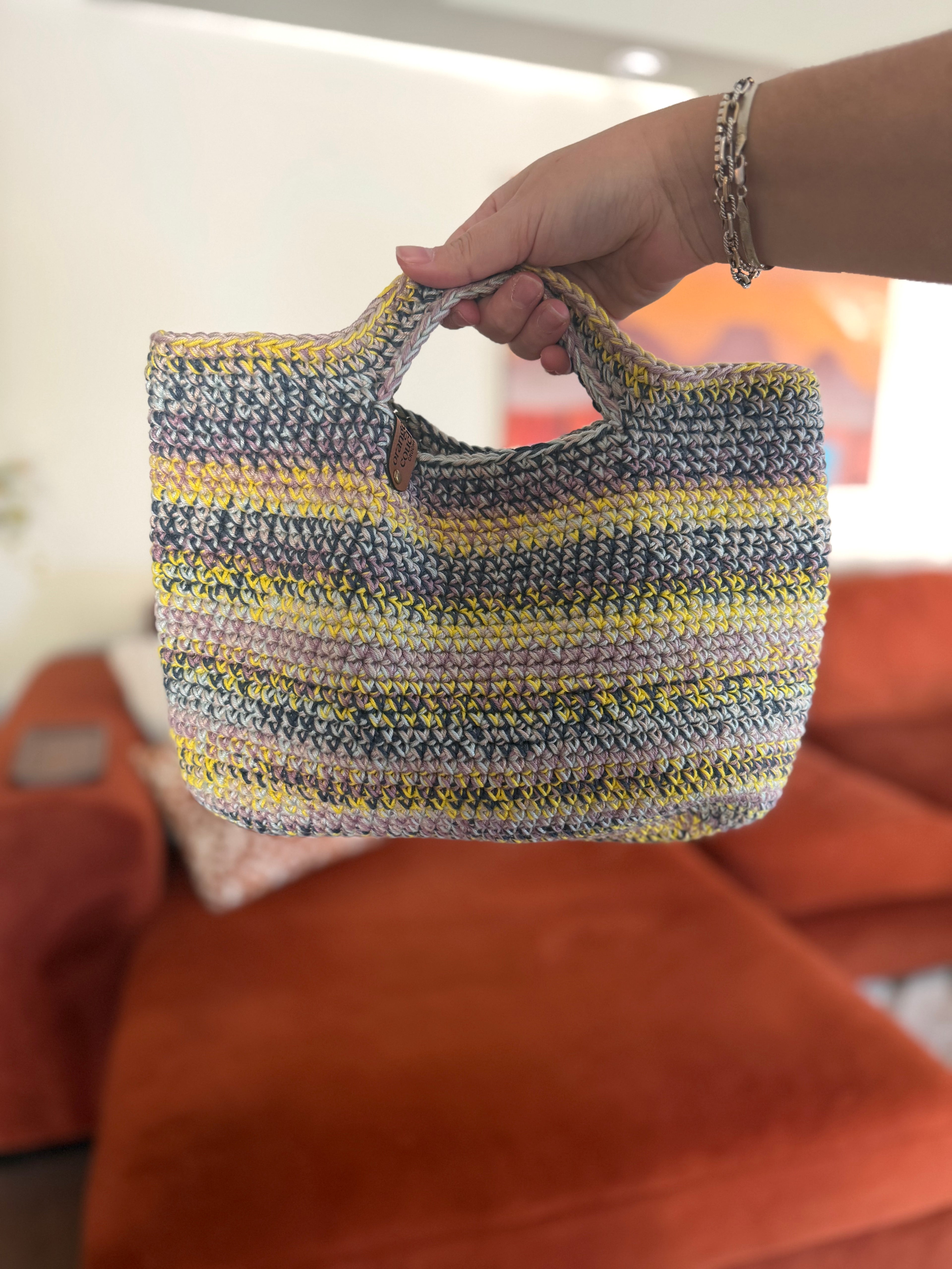 A hand holding a multi-colored small crochet tote bag in front of an orange couch. The bag is dark blue, light, yellow, purple, and beige. 