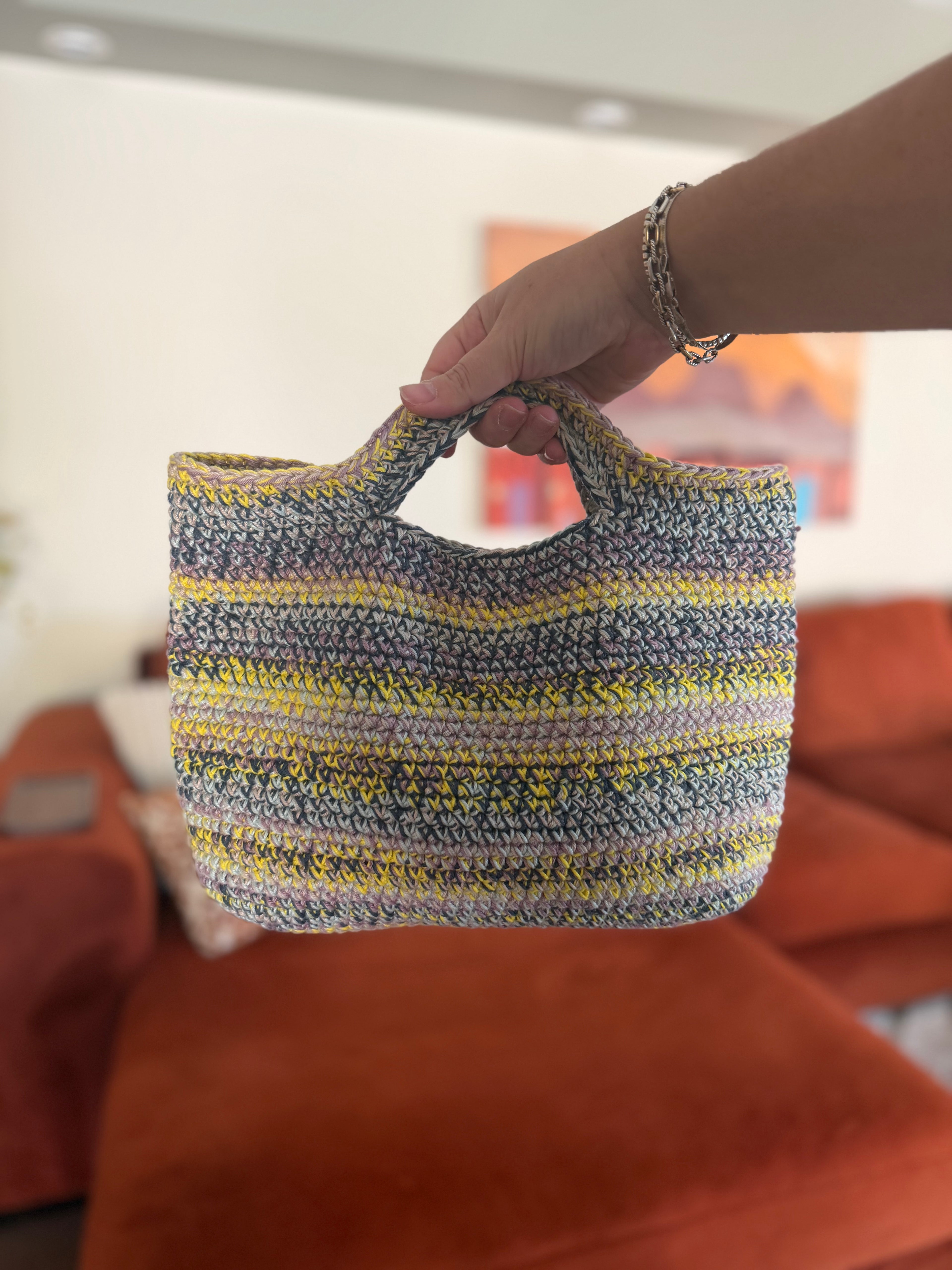 A hand holding a multi-colored small crochet tote bag in front of an orange couch. The bag is dark blue, light, yellow, purple, and beige. 