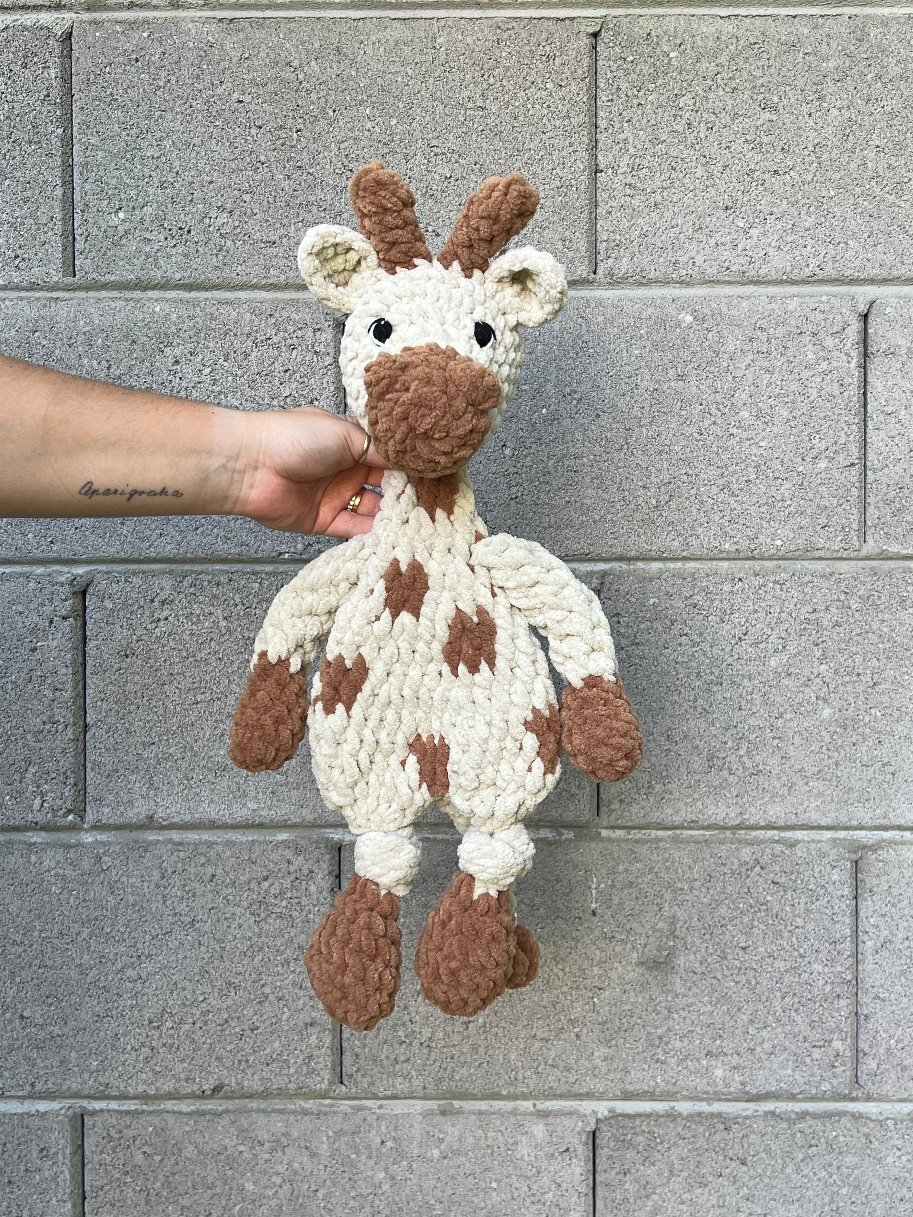 almond and brown crochet giraffe in front of grey brick wall