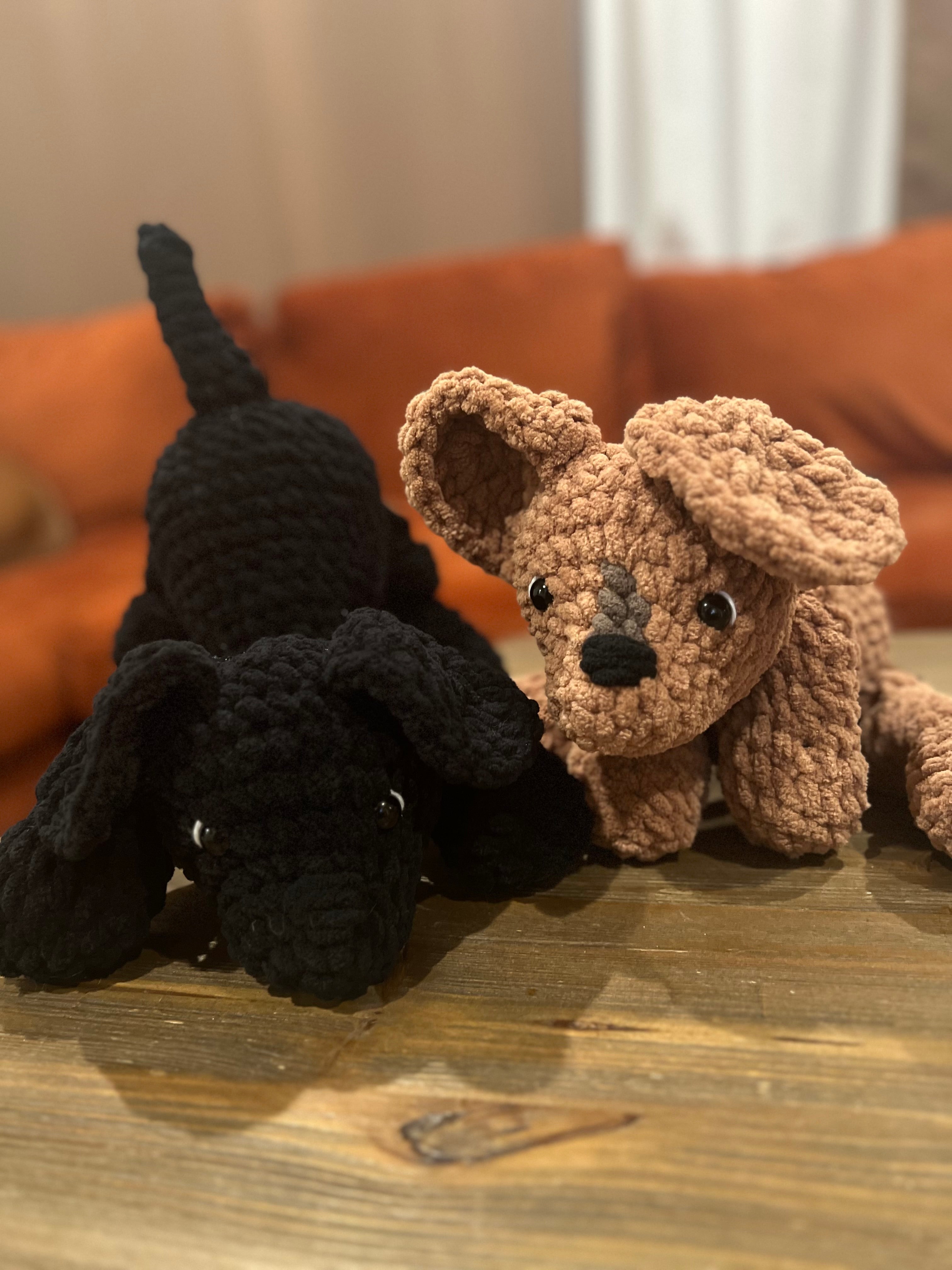 One black crochet dog and one brown crochet dog on wooden table with orange couch in the background